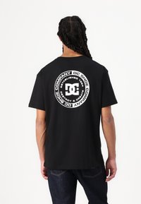 Juodas medvilnės marškinėliai su dideliu baltu apvaliu DC Shoes logotipu nugarinėje pusėje, su užrašu "Įsteigta 1994" ir "Nugriauti ir Kurti."