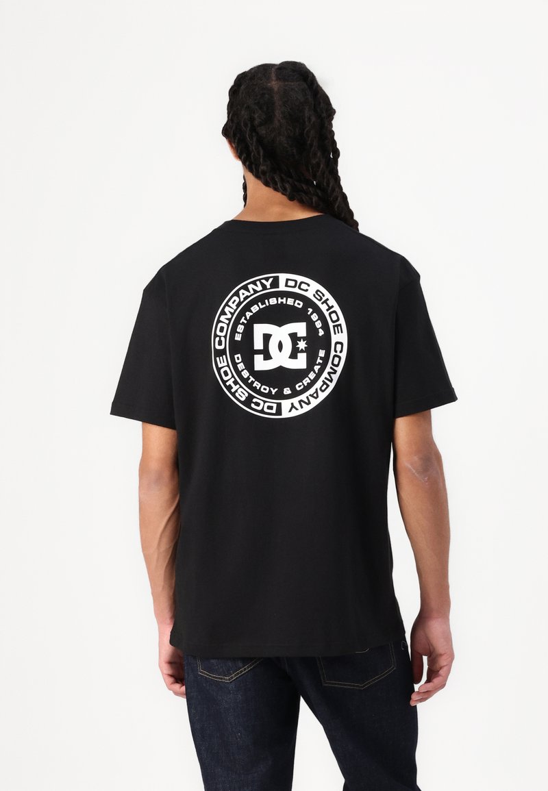 Čierne bavlnené tričko s veľkým kruhovým logom DC Shoes v bielej farbe na chrbte, s textom "Založené 1994" a "Zničiť a vytvoriť."