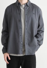 Harmaa napillinen paita pehmeästä kankaasta, jossa klassinen kaulus, vasemmassa rinnassa logo ja rento istuvuus. Päällä vaaleanharmaa paita.