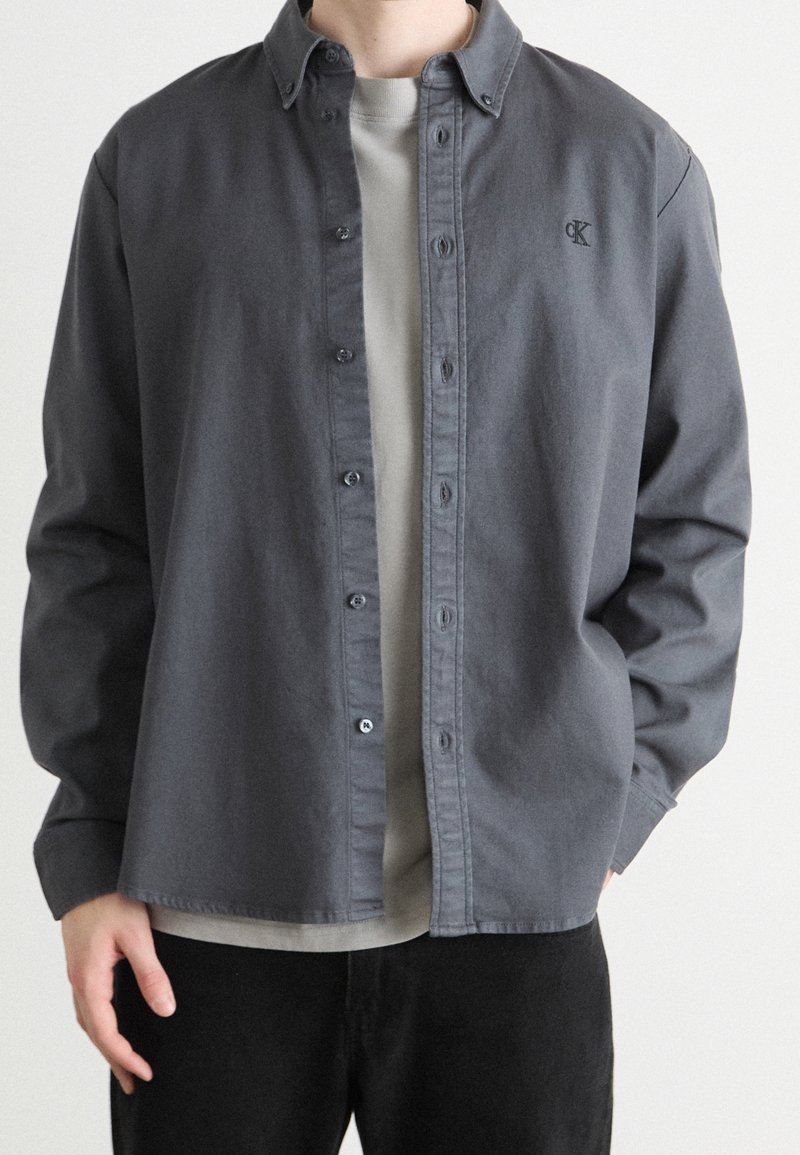 Harmaa napillinen paita pehmeästä kankaasta, jossa klassinen kaulus, vasemmassa rinnassa logo ja rento istuvuus. Päällä vaaleanharmaa paita.