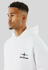 Jonge man met kort krullend haar, gekleed in een witte hoodie met een zwart PXP-logo en een kruisvormige oorbel, kijkend naar links op een grijze achtergrond.