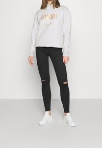 Sudadera gris con "TOMMY" en letras metálicas, combinada con jeans skinny negros rasgados y zapatillas blancas. Diseño sencillo y apariencia casual.