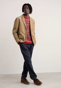 Brūns korduroja žakets, sarkans rakstu džemperis, tumši zilas bikses un brūnas šņorējamas kurpes. Ievērojamas detaļas ietver kabatu un teksturētu dizainu.