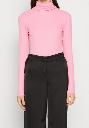 Vrouw draagt een lichtroze coltrui met lange mouwen, ingestopt in zwarte wijde broek met hoge taille.