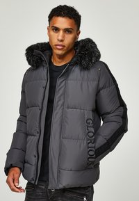 Graue gepolsterte Jacke mit einer Kapuze, gefüttert mit Kunstfell, die das Wort "GLORIOUS" auf der Seite gestickt hat und strukturierte schwarze Streifen an den Ärmeln aufweist.