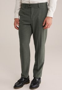 Maßgeschneiderte Hose aus grünem Textilgewebe mit einem flachen Vorderteil, ausgestattet mit seitlichen Taschen und kombiniert mit schwarzen Slippern.