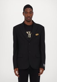 Blazer nero su misura con revers a lancia, due bottoni e tasche laterali. Presenta un badge logo sul petto e un tessuto testurizzato.