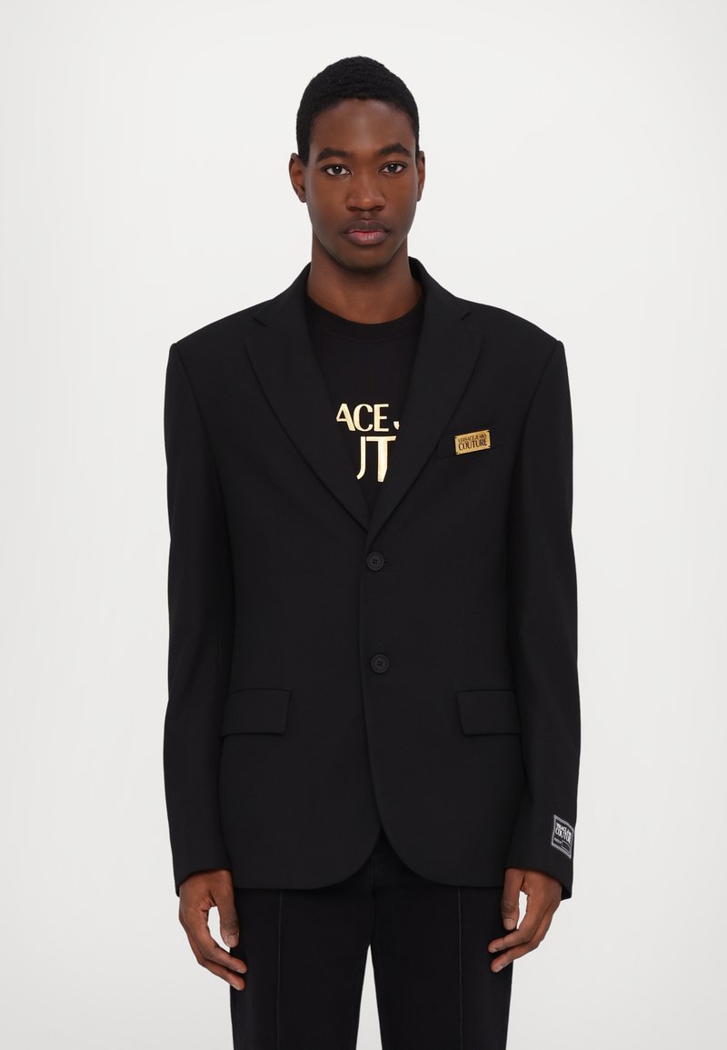 Blazer nero su misura con revers a lancia, due bottoni e tasche laterali. Presenta un badge logo sul petto e un tessuto testurizzato.