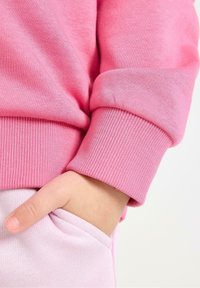 Helles rosa Sweatshirt mit Rippbündchen. Weiche Textur, figurbetonter Schnitt und eine Hand, die in einer passenden hellrosa Tasche ruht.