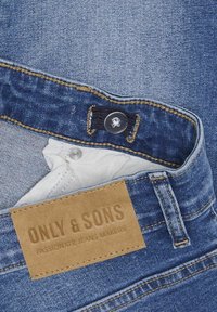 Zili džinsi no zila denima ar gaišu mazgājumu, kuriem ir oranžas dūriens, metāla pogu un brūnu birku ar "ONLY & SONS" zīmola logo.