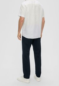 Man die met zijn rug naar de camera staat, gekleed in een wit shirt met korte mouwen, donkerblauwe broek en witte sneakers, tegen een effen lichte achtergrond.