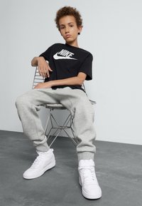Camiseta negra de Nike con logo blanco, pantalones de chándal grises y zapatillas blancas. Cabello rizado, sentado en una silla de alambre contra un fondo claro.