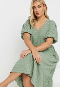 Vihreä ruudullinen mekko, jossa on V-pääntie, puffatut hihat ja virtaava epäsymmetrinen helma, valmistettu kevyestä kankaasta, jossa on tekstuuripinta.