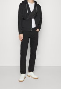 Felpa nera con zip e coulisse, t-shirt bianca, pantaloni neri e sneakers bianche con suola beige. Borsa a tracolla nera.
