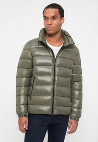 Puffjakke i olivengrøn med lynlås i front, ståkrave og horisontalt quiltet design. Glat, skinnende tekstur med sidelommer.