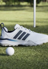 Chaussure de golf blanche Adidas avec des bandes noires sur l'herbe près d'une balle de golf et d'un mât de drapeau en arrière-plan sur un parcours de golf.