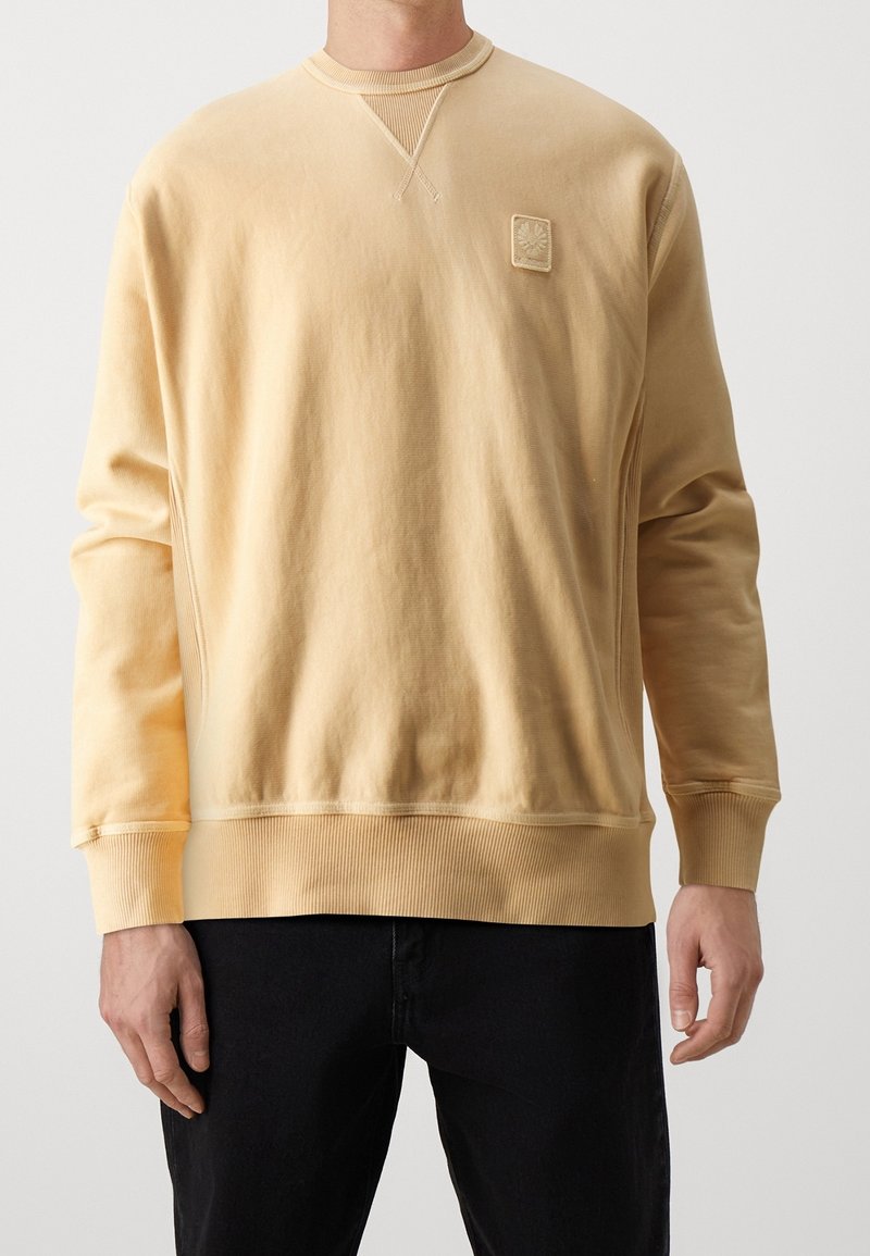 Homme portant un sweat-shirt uni beige clair à manches longues avec poignets et ourlet côtelés, associé à un pantalon noir, debout devant un fond uni.