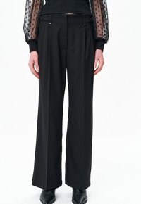 Zwarte wijde broek met een gladde textuur, voorzien van voorplooien en een dubbele knoopsluiting aan de tailleband. Afgeknotte zoom en een nauwsluitende pasvorm.