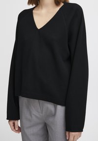 Pull en noir avec texture en gaufre, à col en V et épaules tombantes, assorti à un pantalon tailleur gris clair.