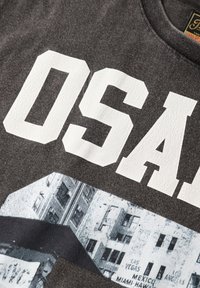Camiseta de algodón gris con grandes letras blancas agrietadas "OSAA" y un gráfico texturizado de un paisaje urbano en blanco y negro debajo.