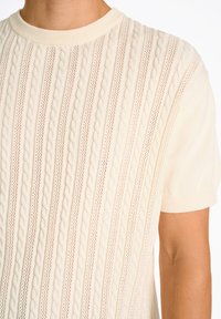 Maglione corto a maniche corte color crema lavorato a maglia, caratterizzato da un motivo a trecce testurizzato con punti aperti e un colletto a girocollo a coste.