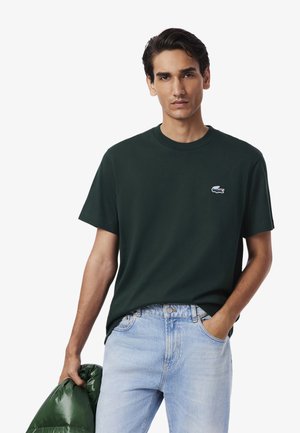 T-shirt verde in cotone con colletto girocollo e maniche corte, caratterizzato da un piccolo logo ricamato blu e bianco sul petto. Indossato con jeans chiari.