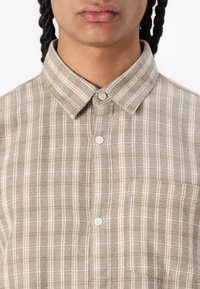 Camicia a quadri marrone chiaro con colletto button-down, caratterizzata da linee beige e bianche, realizzata in tessuto texturizzato. Tre bottoni visibili sulla patta.