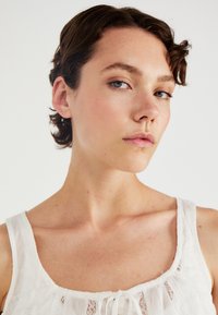 Witte mouwloze top met een kanten rand en strikdetail bij de halslijn. Gladde textuur, zachte stof en een losse pasvorm voor comfort.