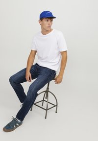 Camiseta de algodón blanca, jeans de denim azul oscuro, gorra azul con texto blanco y zapatillas azules, sentado en un taburete de metal frente a un fondo simple.