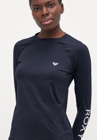 Tamsiai mėlynas ilgomis rankovėmis sportinis marškinėlis su apvaliu kaklu, baltas logotipas ant krūtinės ir "ROXY" užrašas ant rankovės. Sklandžiask, elastingas audinys.