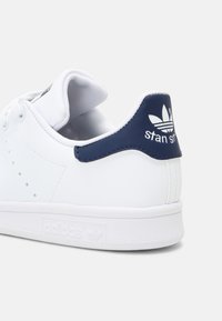 Sneaker bianca in pelle con un tab di colore navy sul tallone. Presenta dettagli perforati, lacci bianchi piatti e una suola bianca testurizzata con logo in rilievo.