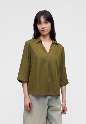 Jonge vrouw met lang donker haar, gekleed in een losse olijfgroene blouse met driekwartmouwen en lichte spijkerbroek tegen een effen achtergrond.