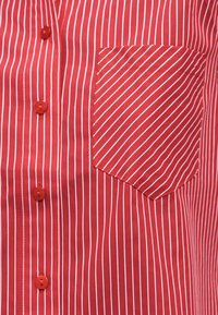 Rdeča srajca z navpičnimi belimi črtami, sprednji žep z diagonalnimi črtami in rdečimi gumbi po sredini.