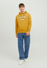 Felpa color giallo senape con tasca a kangaroo frontale e cappuccio con cordino, abbinata a jeans in denim azzurri e sneakers bianche.