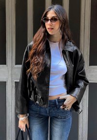 Donna con capelli lunghi castani che indossa occhiali da sole rotondi, giacca di pelle nera, top corto bianco, jeans blu e tiene in mano una borsa nera.