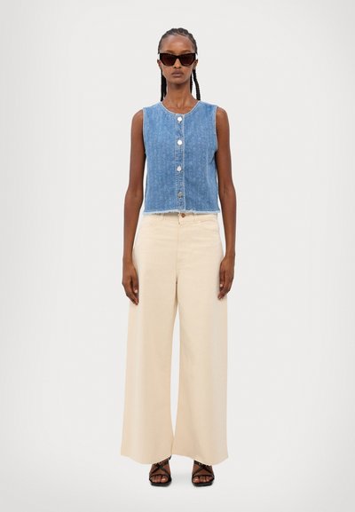 Džínová bezrukávová crop top s bílými knoflíky, světle modrá barva, s třepeným lemem. V kombinaci s béžovými kalhotami s širokými nohavicemi a černými sandály na řemínky.
