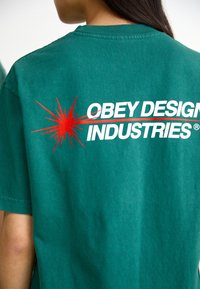 Tricou din bumbac verde, cu un grafic mare roșu care conține textul "OBEY DESIGN INDUSTRIES" și un design de explozie de stea pe spate.