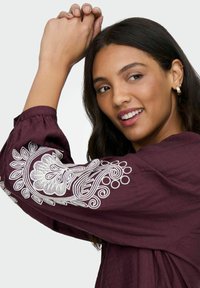 Vinrød broderet bluse med voluminøse ærmer, prydet med indviklede hvide blomster mønstre og tekstureret stof. Model viser designet.
