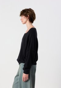 Donna con capelli corti e ricci che indossa un maglione nero largo e jeans azzurri, in piedi di profilo su uno sfondo semplice.