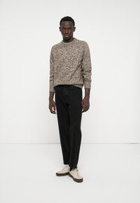 Man wearing beige and brown speckled sweater, black jeans, and beige sneakers, standing against a plain white background.