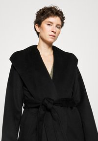 Must värviga pikk mantel laia krae, vööga talje ja tekstuurse kangaga. Omab V-kujulist lõiget rohelise detailiga all.