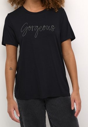 Femme portant un t-shirt noir avec « Gorgeous » écrit en couture blanche cursive sur la poitrine et un pantalon gris foncé.