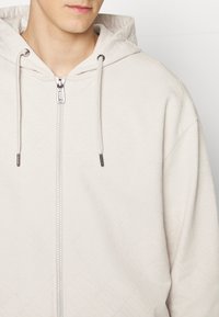 Haut de survêtement zippé beige clair avec un motif texturé. Il comporte une capuche avec cordon de serrage, des éléments en métal argenté et une finition de tissu lisse.