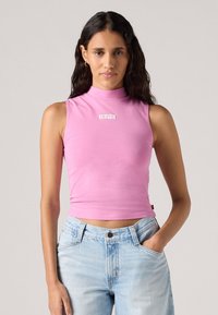 Vaaleanpunainen hihaton mock neck -paita, joka on valmistettu pehmeästä kankaasta, ja keskellä on valkoinen "Levi's" -logo. Täydentää vaaleansiniset farkut.