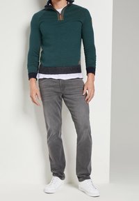 Maglione verde scuro lavorato a maglia con mezza cerniera, orlo inferiore in costine blu navy e jeans grigi aderenti. Completa il look un paio di scarpe da ginnastica bianche.