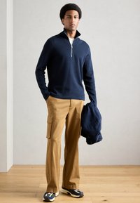 Άνδρας φορώντας ναυτικό μπλέ μισό-φουτερ, καφέ cargo παντελόνια και αθλητικά παπούτσια στέκεται σε ξύλινο δάπεδο κρατώντας μια ναυτική μπουφάν στο χέρι.