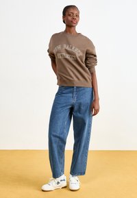 Žena v hnědém svetru "New Balance Athletics", modrých džínách a bílých teniskách stojí na žluté podlaze před bílou zdí.