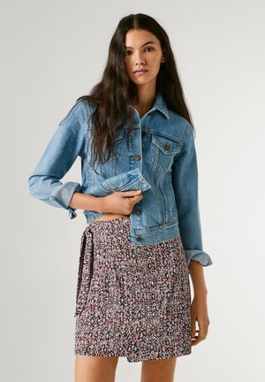 Frau mit langem dunklem Haar, die eine blaue Jeansjacke über einem gemusterten, plissierten Rock trägt und vor einem schlichten hellen Hintergrund steht.