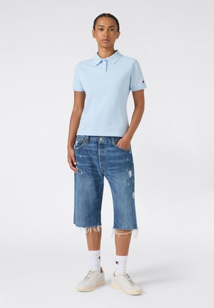 Donna in piedi che indossa una polo azzurro chiaro a maniche corte, pantaloncini in jeans consumati fino al ginocchio, calzini bianchi e scarpe da ginnastica bianche su sfondo bianco.