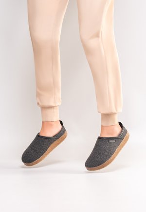 Pieds chaussés de chaussons gris foncé à enfiler avec des semelles en liège, associés à un pantalon jogger beige clair sur un fond uni clair.